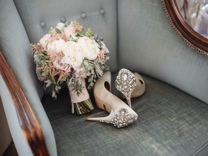 Brides wedding shoes with a bouquet with roses and other flowers on tha arm chair