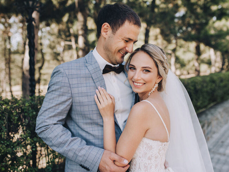 The groom in a gray checkered suit and the blonde bride in a white lace dress tenderly hug in the park on the boulevard. Wedding portrait of newlyweds in love.