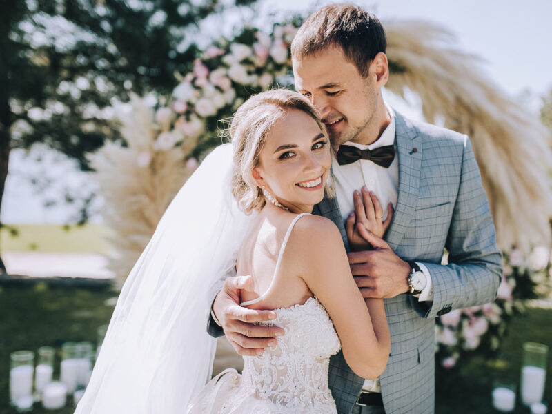 Wedding portrait of smiling newlyweds. Stylish groom in a gray checkered suit and a cute blonde bride in a white lace dress are tenderly hugging in the forest against the background of the arch.
