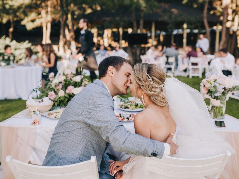 Stylish groom in a gray plaid suit and the blonde bride kiss, sitting at the wedding decorated table. Wedding portrait of newlyweds in love.