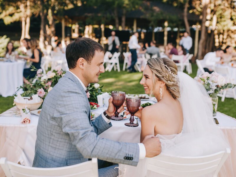 Stylish groom in gray plaid suit and blonde bride sit with faceted glasses of champagne at the wedding decorated table. Wedding portrait of newlyweds in love.