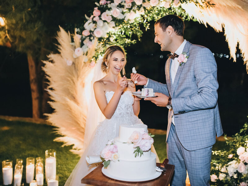 The groom in a gray suit feeds the blonde bride with a spoon with cake on the background of the arch decorated with reeds, flowers, candles. Wedding portrait of laughing newlyweds at night.